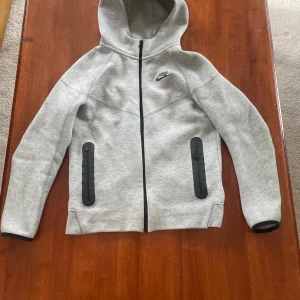 Grå hoodiejacka från Nike, storlek 147- 158 - Snygg grå hoodiejacka från Nike med svart logga på bröstet. Jackan har hel dragkedja framtill, två svarta dragkedjeförsedda fickor och huva. Tillverkad i mjukt material som känns skönt mot huden. Perfekt för chill eller träning.