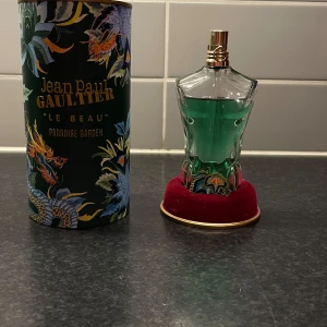 Jean Paul Gaultier Le Beau Paradise Garden - Unik parfym i form av en manlig torso med turkosgrön vätska, guldfärgad spray och detaljerad förpackning med drakmotiv. Flaskan står på en röd sammetsbas med broderad blomma. Perfekt för dig som gillar snygg design och exotiska dofter.