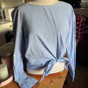 Randig blus med knyte - Snygg blus i ljusblått med vita ränder och långa ärmar. Knyte framtill i midjan och knäppning med knapp i bak. Perfekt för dig som gillar en avslappnad men ändå stilren look. Bomull