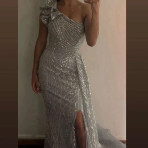 Silverglittrig långklänning oneshoulder - Magisk långklänning i silver med glittriga paljetter och ett unikt mönster. Klänningen har en axel, stor volangdetalj vid axeln och en snygg slits framtill. Perfekt för bal eller fest där du vill sticka ut.