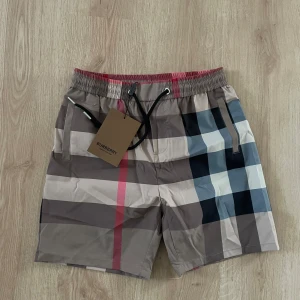 Rutig Burberry shorts med snörning - Snygga shorts från Burberry i klassiskt rutigt mönster med beige, svart, vit och röd. De har elastisk midja med snörning och sidofickor. Tillverkade i bomull och polyester.                                  Helt nya! Skicka bud!                                                        Står L i storlek men sitter mer som S/M    