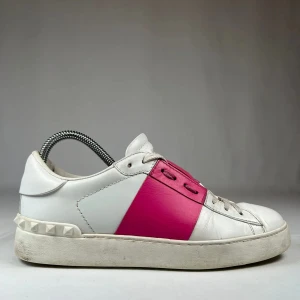Valentino Garavani Open sneakers vit/rosa - Snygga Valentino Garavani Open sneakers i vitt skinn med en bred rosa läderdetalj över mitten och coola nitar på hälen. Klassisk rund tå, snörning och platt sula. Perfekta för dig som vill sticka ut med en lyxig touch.