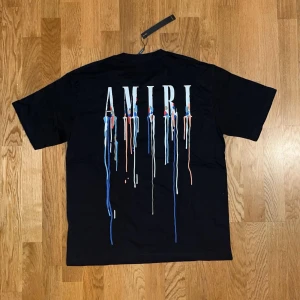 Svart AMIRI t-shirt med drip-print - Svart t-shirt från AMIRI med coolt drip-print i blått, vitt och rött både fram och bak. Oversized fit och klassisk rund hals. Perfekt statement-plagg för dig som gillar streetwear och vill sticka ut.