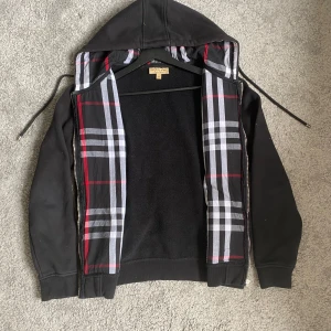 Svart hoodiejacka från Burberry  -  PRIS GÅR ATT ÄNDRAS VID SNABB AFFÄR tar även byten  Svart hoodiejacka från Burberry med klassiskt rutigt foder i huvan i svart, vitt, grått och rött. Jackan har dragkedja, snörning i huvan och två fickor framtill. Tillverkad i mjuk bomull och polyesterblandning, perfekt för en chill och stilren look.