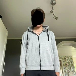 Grå hoodie från Stone Island med dragkedja - Säljer en grå hoodie från Stone Island med svart logotyp-patch på vänster ärm. Tröjan har hel dragkedja framtill, huva med svarta snören och två fickor. Perfekt för dig som gillar streetwear och vill ha en clean look. Pris inte hugget i sten och jag är 172