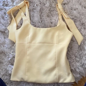 Ljusgul linnetopp med rosettband från ZARA - Supersöt ljusgul linnetopp från ZARA med breda axelband som knyts i rosetter. Toppen har en rak, figurnära passform och är perfekt för varma dagar. Stilren och enkel design som passar till mycket.