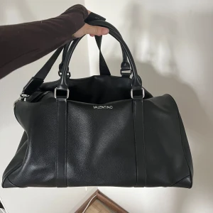 Svart handväska från Valentino - Stilren svart handväska från Valentino i slätt skinn med silverfärgade metalldetaljer. Väskan har dubbla handtag och en avtagbar axelrem, perfekt för dig som vill ha både funktion och snygg design. Rymmer allt du behöver till vardagen och har en klassisk look.