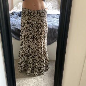Mönstrad långkjol med volang  - Säljer en snygg långkjol med svart och beige mönster och bred smock i midjan. Kjolen har volang nertill som ger en flowy känsla och passar perfekt till sneakers eller sandaler. Superbekväm och lätt att styla till olika toppar.