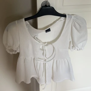 Vit blus med puffärm och knyt - Supersöt vit blus från envii med korta puffärmar och knyt framtill. Blusen har en lätt rynkad struktur, volang nedtill och en luftig känsla. Perfekt för dig som gillar romantisk och trendig stil.