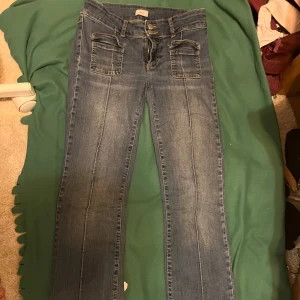 Blå bootcut jeans med fickdetaljer - Snygga blå jeans med bootcut passform och markerade sömmar längs benen. Framtill finns två stora fickor med lock som ger en cool look. Jeansen har klassisk dragkedja och knappstängning. Storlek 38