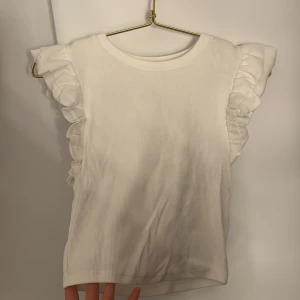 Vit volangtopp från Zara - Supersöt vit topp från Zara med ribbad struktur och volangdetaljer på axlarna. Ärmlös modell som ger en fräsch och trendig look. Perfekt att styla med jeans eller kjol för en cool vibe. Helt oanvänd fick i present kom aldrig till användning 