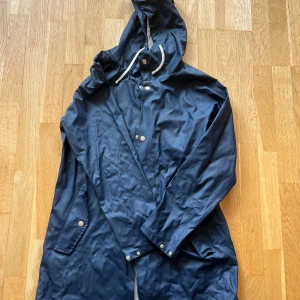 Mörkblå regnjacka  - Snygg mörkblå regnjacka från 157 med huva och vita dragsnören. Jackan har silvriga knappar, stora fickor och är i ett galonliknande material. Jag säljer även en likadan jacka fast i svart om det är av intresse. Hör av dig om du har frågor! 😊