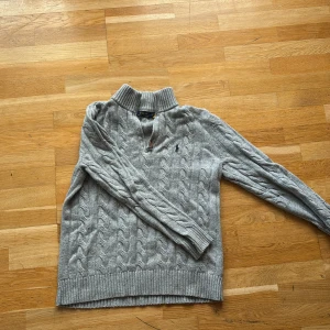 Grå kabelstickad tröja Polo Ralph Lauren - Kabelstickad grå tröja från Polo Ralph Lauren med halv dragkedja och hög krage. Klassisk broderad logga på bröstet och ribbade muddar. Perfekt för lager-på-lager och chill höst- eller vinterstil.