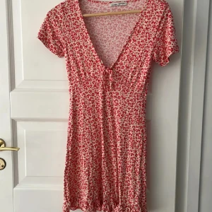 Rödblommig kortklänning med v-ringning - Supersöt kortklänning från Samsøe Samsøe med rött blommönster på vit botten, djup v-ringning och knyt framtill. Klänningen har korta ärmar och volangkant nertill. Perfekt för varma dagar och somriga vibes!
