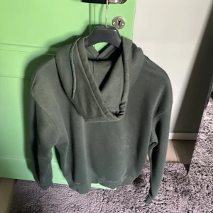 Mörkgrön hoodie med huva och ficka - Säljer en mörkgrön hoodie med klassisk känguruficka och snörning i huvan. Tröjan har lång ärm och ribbade muddar vid ärmslut och nederkant. Perfekt för chill dagar eller när du vill ha en enkel och stilren look.