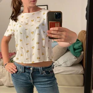 Vit blus med citronmönster - Superfin vit blus med sött citronmönster i gult och svart. Modellen är croppad med korta ärmar och rund halsringning. Perfekt för dig som gillar fräscha och lekfulla prints. Tunn och luftig, passar perfekt till jeans eller kjol.
