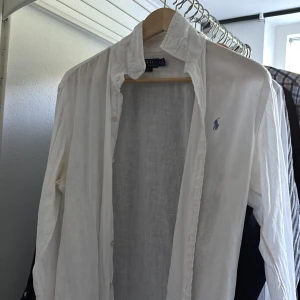 Vit linneskjorta från Polo Ralph Lauren - Klassisk vit skjorta i linne från Polo Ralph Lauren med blå broderad logga på bröstet. Skjortan har lång ärm, knappar framtill och en avslappnad passform. Perfekt för en stilren och fräsch look.
