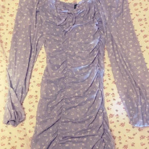 Ljusblå blommig klänning - Supersöt ljusblå klänning från H&M med små vita blommor, långa genomskinliga ärmar och snygg rynkdetalj framtill. Har en rosett vid bröstet som man kan se på bilden , lätt material som passar perfekt till vår och sommar. Använd endast en gång. 