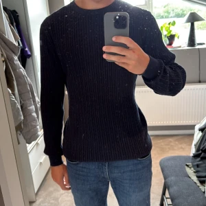 Svart ribbstickad tröja - Säljer en svart ribbstickad tröja med rund hals och långa ärmar. Tröjan har en klassisk och stilren look som passar perfekt till jeans eller chinos. Materialet är mjukt och ger en skön känsla, perfekt för kyligare dagar.