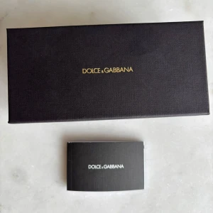 Svarta solglasögon Dolce & Gabbana - Snygga solglasögon från Dolce & Gabbana med fyrkantiga svarta bågar och guldiga skalmar. Glasen är mörktonade och ramen har en exklusiv känsla. Kommer med originalfodral och box. Perfekt accessoar för en trendig look. Originalpris: 3500. Box kommer med