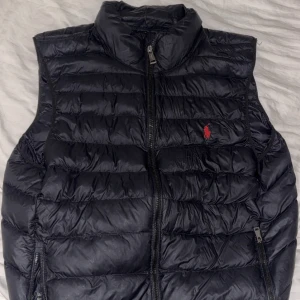 Svart dunväst från Polo Ralph Lauren - Snygg svart dunväst från Polo Ralph Lauren med klassisk röd logga på bröstet. Västen har hög krage, två dragkedjefickor och quiltad design. Högra dragkedjan funkar ej men annars e allt som de ska. 