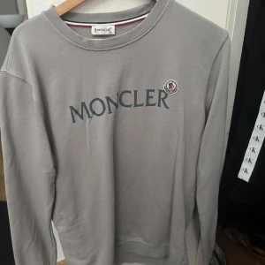 Grå Moncler sweatshirt XL - Säljer en stilren grå sweatshirt från Moncler i storlek XL. Tröjan har rund hals, lång ärm och klassisk Moncler-logga på bröstet samt tryckt Moncler-text framtill. Perfekt för dig som gillar exklusiva och enkla plagg. Passar mest L 