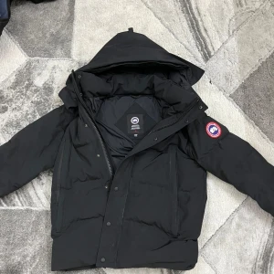 Svart dunjacka från Canada Goose - Säljer en svart dunjacka från Canada Goose med avtagbar huva . Jackan har en klassisk design med dragkedja och knappar framtill samt en ikonisk logga på ärmen. Perfekt för kalla vinterdagar. 