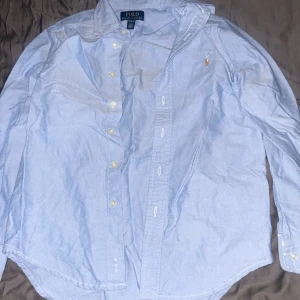 Ljusblå skjorta från Polo Ralph Lauren - Klassisk ljusblå skjorta från Polo Ralph Lauren med broderad logga på bröstet. Skjortan har lång ärm, knappar framtill och är tillverkad i bomull. Perfekt för en clean och stilren look.