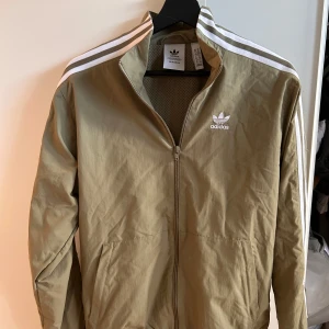 Olivgrön vindjacka från Adidas XS - Snygg olivgrön vindjacka från Adidas med klassiska vita ränder längs ärmarna och broderad logga på bröstet. Jackan har dragkedja framtill, ståkrage och meshfoder. Perfekt för en sportig och avslappnad stil.
