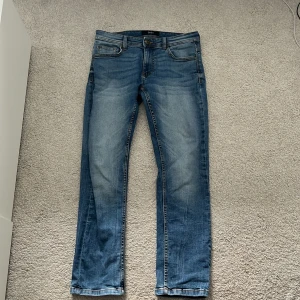 Blå jeans från SMOG, storlek 30/30 - Snygga blå jeans från SMOG i klassisk femficksmodell. Jeansen har en rak passform med lätt slitning på låren och kontrastsömmar. Tillverkade i bomull för en skön känsla och passar perfekt till en avslappnad stil.