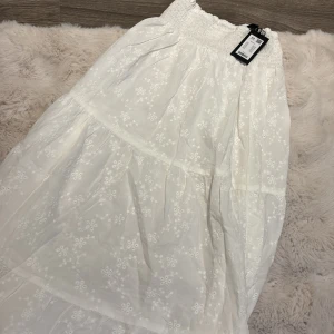 Vit långkjol med broderade blommor - Superfin vit långkjol från Blossom med broderade blommor och smock i midjan. Kjolen har volanger och är gjord i ett lätt bomullstyg som känns luftigt och somrigt. Perfekt för dig som gillar romantisk och söt stil.