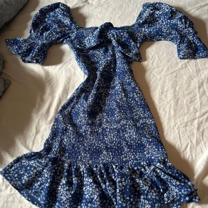 Blå blommig kortklänning Gina Tricot - Supersöt blå kortklänning från Gina Tricot med vita blommor, volangkant nertill och puffiga ärmar. Klänningen har knyt fram och smockad midja för en snygg passform. Perfekt för sommarens alla tillfällen!