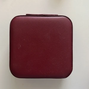 Vinröd smyckesväska i skinnimitation - Snygg och kompakt vinröd smyckesväska i skinnimitation med dragkedja. Insidan är ljusbeige och har flera fack och rullar för ringar, samt krokar och ficka i locket. Perfekt för att hålla koll på dina smycken när du är på språng.