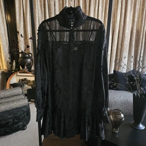 Custommade klänning- Elorie - Supersnygg svart kortklänning med broderade blommor, spetsdetaljer och volangkant. Hög krage med dekorativa knappar och transparenta partier upptill. Långa ärmar med manschett och knapp. Perfekt för dig som gillar unika detaljer och romantisk stil. (Prislapp saknas, men är aldrig använd)