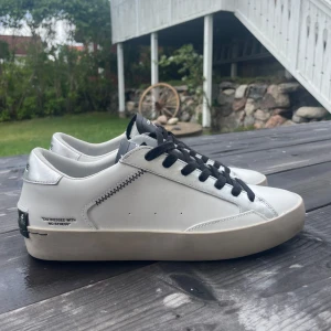 Vita sneakers från Crime London strl 41 - Snygga vita sneakers från Crime London med svarta snören och detaljer. Skorna är i skinn och har en klassisk låg profil med svart logga på plösen och hälen. Sulan är vit med svart undersida och cool textdetalj på sidan. Perfekta för en clean och modern stil. Helt oanvända, nypris 1900kr