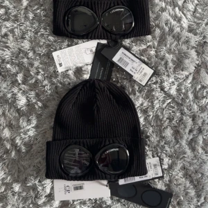 Svart C.P. Company goggle-mössa - Svart ribbstickad mössa från C.P. Company med ikoniska goggle-linser framtill på uppviket. Tillverkad i mjuk bomull och har en clean, urban look. Perfekt accessoar för dig som vill sticka ut med streetwear-vibe. 1190kr/st eller köp båda för 2k!!