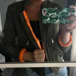 Olivgrön bomberjacka från DM retro - Snygg olivgrön bomberjacka från DM retro med orange insida, finns inte att få tag på längre då företaget slutat sälja den. Jackan har dragkedja framtill och på ärmen, samt ribbade muddar vid ärmslut och nederkant. Perfekt nu inför våren🌸💚