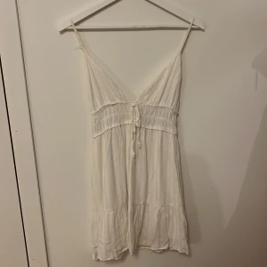 Vit klänning med smala axelband - Söt vit klänning med smala axelband och v-ringning. Den har en knytning i midjan och en lätt volangdetalj vid fållen. Perfekt för varma sommardagar eller en avslappnad dag på stranden.