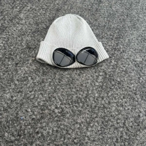 Grå mössa med glasögondetalj från C.P. Company - Snygg grå ribbad mössa från C.P. Company med unika glasögondetaljer framtill i bra skick. Perfekt för att ge din outfit en cool touch. Tillverkad i mjukt material för extra komfort. Hör av dig vid frågor eller funderingar ✅