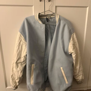 Ljusblå och vit bomberjacka från H&M - Snygg ljusblå och vit bomberjacka från H&M med knappar framtill. Jackan har långa ärmar och ribbade muddar vid ärmslut och nederkant. Perfekt för en cool och avslappnad stil.