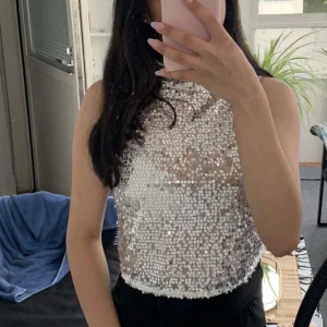 Paljettopp från Gina Tricot - Säljer en glittrig paljettopp från Gina Tricot i silver. Toppen är ärmlös och perfekt för festliga tillfällen. Den har en rund halsringning och knäppning i nacken. Passar utmärkt för att lysa upp kvällen! ✨Ny utan prislapp.