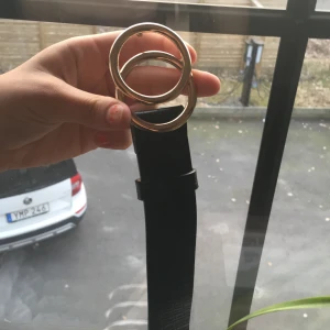 Svart bälte med guldfärgat spänne - Snyggt svart bälte med ett stilrent guldfärgat spänne i dubbel ringdesign. Bältet har en texturerad yta som ger en extra touch av elegans. Perfekt för att lyfta vilken outfit som helst.
