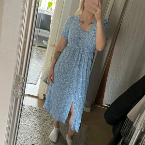 Blå blommig klänning - Blå klänning med blommigt mönster och v-ringning. Klänningen har korta ärmar, pärlemor knappar framtill och resår i ryggen. Snygg och skön passform.  Perfekt för en somrig look med sin luftiga design och slitsdetalj. 100% viskos. Strl S. Ord pris 699:- 