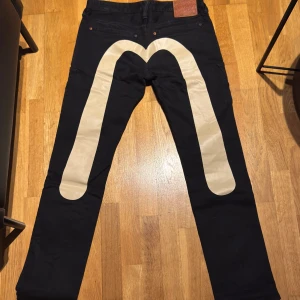 Svarta jeans från Evisu med vit logga - Säljer ett par svarta jeans från Evisu med en stor vit logga på baksidan. Jeansen har en klassisk femficksdesign och en läderpatch med Evisus ikoniska logga. Perfekta för dig som vill ha något unikt i garderoben.