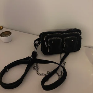 Svart axelväska med kedjedetalj - Snygg svart axelväska med flera dragkedjefickor framtill och en cool kedjedetalj på axelremmen. Perfekt för att hålla dina saker organiserade och ge en edgy touch till din outfit.