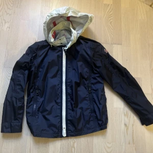 Mörkblå vindjacka från Moncler - Säljer en stilren mörkblå vindjacka från Moncler Grenoble. Jackan har en vit dragkedja och en justerbar huva. Perfekt för blåsiga dagar och har en snygg logga på insidan. Passar bra för både stad och natur.