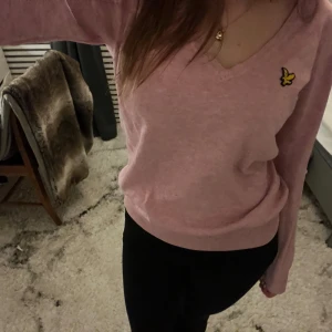 Rosa tröja från Lyle & Scott - Säljer en rosa tröja från Lyle & Scott med v-ringning och ett litet gult emblem på bröstet. Tröjan har långa ärmar och är perfekt för en avslappnad stil. Passar bra till både jeans och kjol. 💖