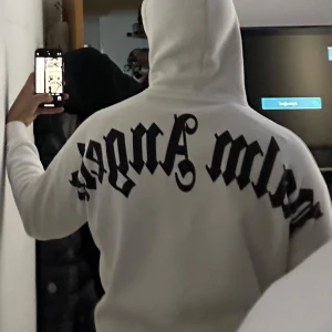 Vit hoodie från Palm Angels - Säljer en stilren vit hoodie från Palm Angels med svart text på både fram- och baksidan. Hoodien har en klassisk passform med en stor ficka framtill och justerbar huva. Perfekt för en avslappnad look.
