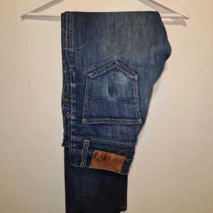 Blå jeansbyxor från Tiger of Sweden  - Snygga blå jeans med klassisk femficksdesign och slitningar för en trendig look. Perfekta för en avslappnad stil. Lite slitning av snusdosa därav lägre pris.