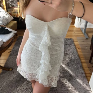 Vit axelbandslös klänning med volang - Säljer en söt vit axelbandslös klänning med volangdetalj framtill. Klänningen är kort och har spetsdetaljer. Perfekt för studenten!